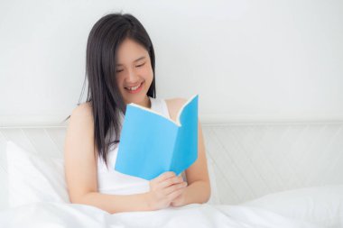 Genç Asyalı kadın rahatça kitap okuyor. Yatak odasındaki yatakta rahat rahat uyuyor. Kadın boş vakitlerinde kitap okuyor..