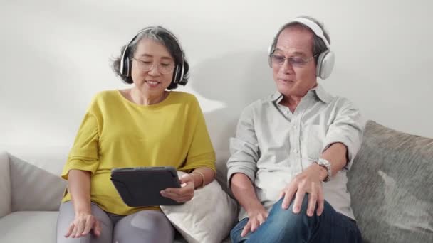 快乐的老夫妇们 戴着耳机 在家里客厅里听音乐 看社交媒体的平板电脑 老年男女们放松舒适地享受着生活方式的概念 — 图库视频影像
