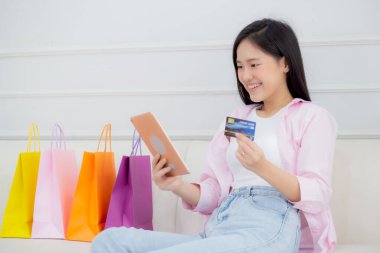 Young asian woman sitting on sofa using digital tablet shopping online with credit card while paper bag on couch, female paying with transaction financial, purchase and payment, business concept.
