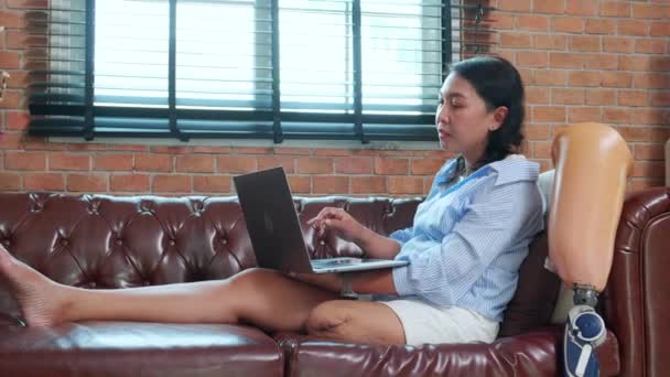 若いアジアの女性は 自宅のリビングルームに義足のラップトップコンピュータを使用してソファーに座って障害 楽観的な ライフスタイルと障害を持つノートブックと義肢に取り組む女性 — ストック動画