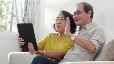 Mutlu son sınıf çifti koltukta oturup tablet kullanırken, video evde aile ile çevrimiçi arama yaparken, mutlu yaşlı erkek ve kadın aile ile uzak ve keyifli bir şekilde sohbet ediyorlar..