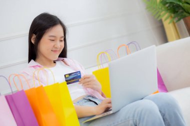 Kanepede oturan Asyalı genç bir kadın internet üzerinden dizüstü bilgisayar alışverişi yapıyor. İnternete kredi kartı alıyor, alışveriş, alım-satım ve ödeme ile ödeme yapan mutlu bir kız..