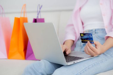 Closeup hands of woman sitting on sofa using laptop computer shopping online with credit card while paper bag on sofa, female paying with transaction financial, purchase and payment, business concept.