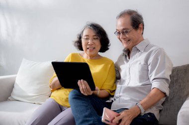 Mutlu son sınıf çifti koltukta oturup tablet kullanırken, video evde aile ile çevrimiçi arama yaparken, mutlu yaşlı erkek ve kadın aile ile uzak ve keyifli bir şekilde sohbet ediyorlar..