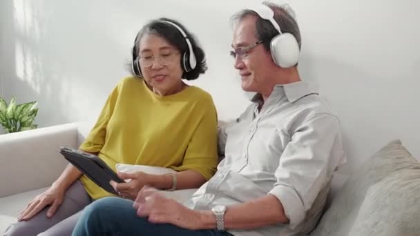 ハッピーファミリーシニアカップルは 音楽を聴いたヘッドフォンを着て 高齢者 女性のリビングルームでソーシャルメディアにタブレットを見ながら 楽しく ライフスタイルのコンセプトでリラックスして居心地の良い — ストック動画