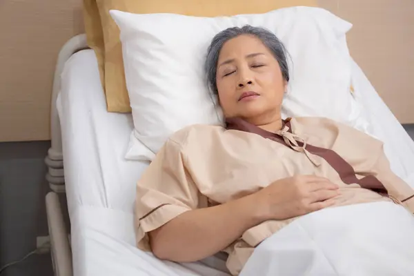 Asian elderly woman asleep on bed with surveillance in hospital ward, patient senior unconscious lying on bed, elder ill while relax at clinic, ailment and treatment, medical and insurance concept.