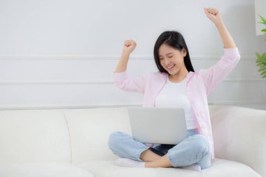 Mutlu Asyalı genç bir kadın koltuğunda oturup dizüstü bilgisayarda çalışırken evde başarılı ve heyecanlı, not defteri ve başarıyı kullanan serbest bir kadın, iş ve yaşam tarzı konsepti.