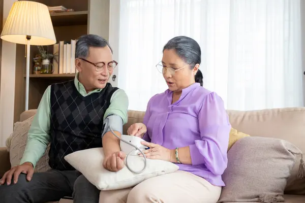 Asian senior couple sitting on sofa checking blood pressure with pressure gauge in living room at home, elderly man and woman sitting on couch checkup health and pressure, medical of patient.