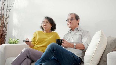 Mutlu Asyalı yaşlı çift kanepede oturup rahatça televizyon izliyor ve evde oturma odasında eğleniyor. Yaşlı bir adam ve kadınla birlikte mutlu bir aile hafta sonu televizyon seyrediyor..