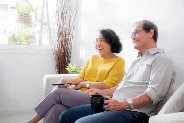 Happiness asian senior couple sitting on sofa watching TV with relax and enjoy in living room at home, happy family with elderly man and woman watching television, entertainment in weekend.