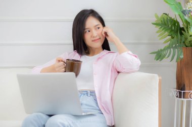 Young asian business woman work from home with laptop computer and drinking coffee on sofa in living room, freelance girl using notebook sitting on couch with comfort and relax, lifestyles concept.