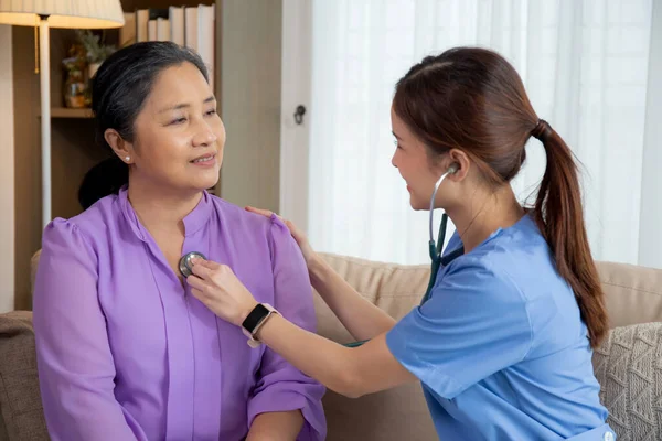 Young asian nurse or caregiver sitting on sofa holding stethoscope listen heartbeat with senior woman in living room at home, doctor check cardiologist disease with elderly, medical and insurance.