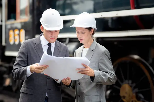 Young caucasian engineer man and woman in suit checking train looking blueprint in station, team engineer inspect system transport, technician examining infrastructure, transportation and industry.