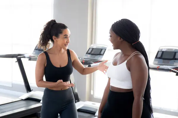 Two young woman talking in the gym, woman in sportswear and friend or coach relax and discussing while break in fitness, workout and exercise for health, diversity and ethnicity, sport concept.