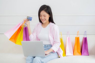 Kanepede oturan Asyalı genç bir kadın bilgisayarla online alışveriş yapıyor. Kredi kartıyla ve kesekağıdıyla ödeme yapıyor. İşletme konseptiyle ödeme yapan mutlu bir kadın..