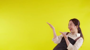 Önlük giymiş Asyalı genç bir baristanın portresi sarı arka planda heyecanlı bir şekilde sunuluyor, garson ya da girişimci neşeli ve bir şeyler gösteriyor, küçük işletme ya da girişim, kafe garsonu..