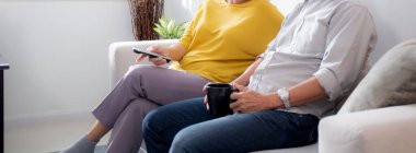 Mutlu Asyalı yaşlı çift kanepede oturup rahatça televizyon izliyor ve evde oturma odasında eğleniyor. Yaşlı bir adam ve kadınla birlikte mutlu bir aile hafta sonu televizyon seyrediyor..