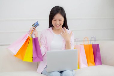Kanepede oturan Asyalı genç bir kadın bilgisayarla online alışveriş yapıyor. Kredi kartıyla ve kesekağıdıyla ödeme yapıyor. İşletme konseptiyle ödeme yapan mutlu bir kadın..