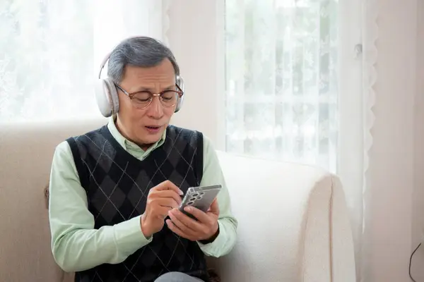 Asian senior man wearing headphone listening music with smartphone sitting on sofa for relax in living room at home, elderly wearing headset listening radio on smart phone, lifestyles concept.