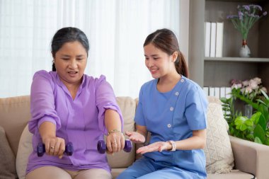 Evde oturma odasında terapi ve rehabilitasyon görürken, bakıcı ya da fizyoterapist yaşlılara yardım ederken, son sınıf öğrencisi olan Asyalı bir kadın ya da hemşire kol egzersizi için halteri kaldırıyor..