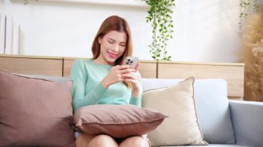 Genç Asyalı mutlu bir kadın kanepede oturmuş gülümsüyor. Oturma odasında rahat bir şekilde akıllı telefon kullanıyor. Kanepede oturmuş sosyal medyayı seyrediyor..