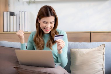 Kanepede oturan Asyalı genç bir kadın internet üzerinden dizüstü bilgisayar alışverişi yapıyor. Kredi kartıyla internete alışveriş yapıyor. Mutlu olurken, kadın ödemesi kanepe, satın alma ve ödeme, iş konsepti..