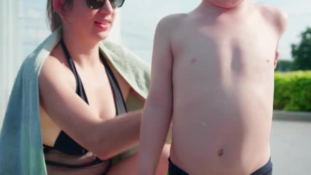 在阳光充足的日子里 白种人妈妈在游泳池里给儿子的后背涂上防晒霜 妈妈在游泳池边涂上乳液或防晒霜 以保护男孩的皮肤不受紫外线感染 并保护他们的家人和皮肤 — 图库视频影像