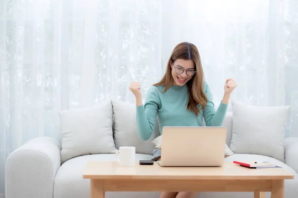 Young asian woman wearing glasses sitting sofa working on laptop computer in living room at home, woman work from home with telework, freelance working and success, business and digital life.