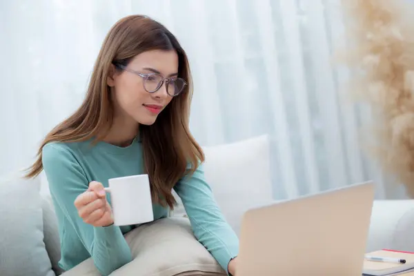 Young asian woman wearing glasses sitting sofa working on laptop computer and drinking coffee in living room at home, woman work from home with telework, freelance working, business and digital life.