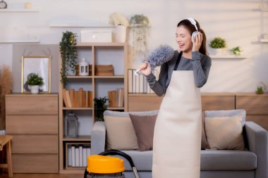 Genç Asyalı bir kadın, kulaklıkla müzik dinlerken, oturma odasında temizlik yaparken, ev işi yaparken, evde toz alırken, şarkı söylerken, ev işi yaparken, yaşam tarzı konsepti.