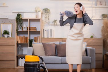 Genç Asyalı bir kadın, kulaklıkla müzik dinlerken, oturma odasında temizlik yaparken, ev işi yaparken, evde toz alırken, şarkı söylerken, ev işi yaparken, yaşam tarzı konsepti.