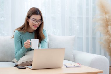 Genç Asyalı kadın gözlüklü koltukta oturuyor dizüstü bilgisayarda çalışıyor ve evdeki oturma odasında kahve içiyor, kadın evden telgrafla çalışıyor, serbest çalışıyor, iş ve dijital hayatla uğraşıyor..