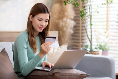 Kanepede oturan Asyalı genç bir kadın internet üzerinden dizüstü bilgisayar alışverişi yapıyor. İnternete kredi kartı alışverişi yapıyor. Kanepe, satın alma ve ödeme, iş ve yaşam tarzları ile kadın ödemesi yapıyor..