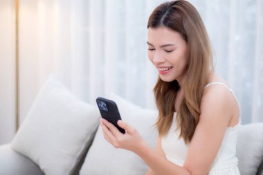 Kanepede oturan Asyalı genç bir kadın evdeki oturma odasında akıllı telefon kullanıyor, sosyal medyayı telefonda sohbet için kullanıyor ve rahatlama, yaşam tarzı konsepti olan uzak sohbetler yapıyor..