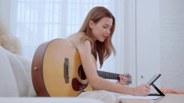 Genç Asyalı bir kadın kanepeye oturup pratik yapmak için oturma odasında gitar çalıyor. İnternetten müzik ve gitar çalmayı öğrenme konsepti hakkında ders izliyor..