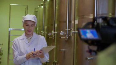 Şarap üreticisi kadın şarap üretimini kontrol ediyor ve inceliyor ve fabrikada kamerayla sunuyor, müfettiş kaliteyi kontrol ediyor ve şarap deposunu depoda mayalıyor, endüstriyel konsept.
