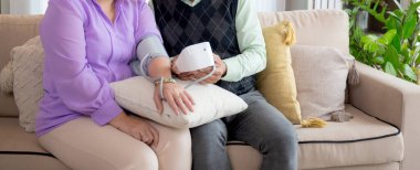 Kanepede oturan Asyalı yaşlı çift oturma odasındaki basınç göstergesiyle kan basıncını ölçüyor, yaşlı adam ve kadın kanepede oturuyor sağlık ve basınç kontrolü yapıyor,.