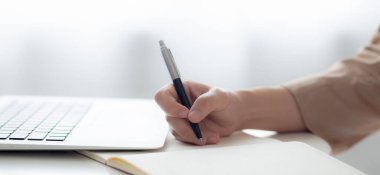 Yakın çekimde genç Asyalı kadın dizüstü bilgisayarı evde çalışırken ve kullanırken planlamak için deftere yazı yazıyor. Finans, kadın çalışmaları, iş ve iletişim konsepti hakkında kadın notları..