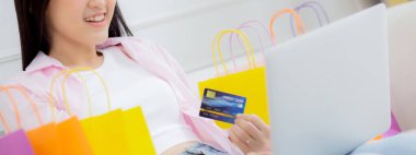 Young asian woman sitting on sofa using laptop computer shopping online with credit card buying to internet, happiness girl paying with transaction financial, purchase and payment, business concept.