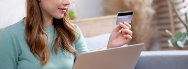 Kanepede oturan Asyalı genç bir kadın internet üzerinden dizüstü bilgisayar alışverişi yapıyor. İnternete kredi kartı alışverişi yapıyor. Kanepe, satın alma ve ödeme, iş ve yaşam tarzları ile kadın ödemesi yapıyor..