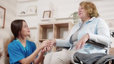 Bakıcı Asyalı kadın tekerlekli sandalyede oturan yaşlılara bakıyor, evdeki oturma odasında cesaretlendirme, son sınıf terapisi ve doktorla rehabilitasyon, rehabilitasyon, sigorta ve tıbbi tedavi için teselli buluyor..
