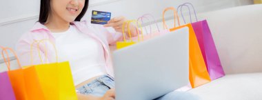 Kanepede oturan Asyalı genç bir kadın internet üzerinden dizüstü bilgisayar alışverişi yapıyor. İnternete kredi kartı alıyor, alışveriş, alım-satım ve ödeme ile ödeme yapan mutlu bir kız..
