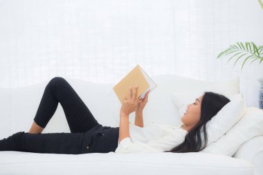 Kanepede uzanmış kitap okuyan genç Asyalı bir kadın evinde dinleniyor. Hafta sonu rahat, hobi ve eğlence dolu bir kanepede edebiyat okuyan bir kadın..