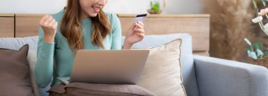 Kanepede oturan Asyalı genç bir kadın internet üzerinden dizüstü bilgisayar alışverişi yapıyor. Kredi kartıyla internete alışveriş yapıyor. Mutlu olurken, kadın ödemesi kanepe, satın alma ve ödeme, iş konsepti..