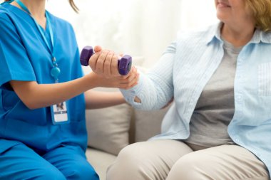 Evde oturma odasında terapi ve rehabilitasyon görürken, bakıcı ya da fizyoterapist yaşlılara yardım ederken, son sınıf öğrencisi olan Asyalı bir kadın ya da hemşire kol egzersizi için halteri kaldırıyor..