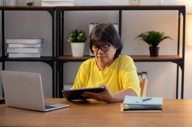 Üst düzey Asyalı kadın tablet okuyup ders çalışırken dizüstü bilgisayarı evde oturma odasında, yaşlı kadın konsantrasyon, yaşam tarzı ve eğitimle eğitimi öğrenirken,.