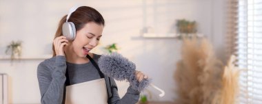 Genç Asyalı bir kadın, kulaklıkla müzik dinlerken, oturma odasında temizlik yaparken, ev işi yaparken, evde toz alırken, şarkı söylerken, ev işi yaparken, yaşam tarzı konsepti.