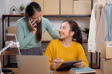 Tablet üzerinde çalışan iki genç Asyalı kadın çevrimiçi iş yaparken ve planlarken ve evde akıllı telefonla konuşarak siparişi kontrol ederken, çevrimiçi satıcı, girişimci ve kargo şirketiyle perakende satış yaparken,.