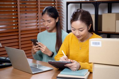 Tablet üzerinde çalışan iki genç Asyalı kadın çevrimiçi iş yaparken ve planlarken ve akıllı telefondaki konuşmayla siparişleri kontrol ederken, çevrimiçi satıcı, girişim şirketi ve kargoyla perakende satış yaparken.