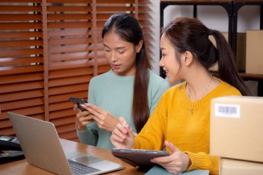 Tablet üzerinde çalışan iki genç Asyalı kadın çevrimiçi iş yaparken ve planlarken ve akıllı telefondaki konuşmayla siparişleri kontrol ederken, çevrimiçi satıcı, girişim şirketi ve kargoyla perakende satış yaparken.